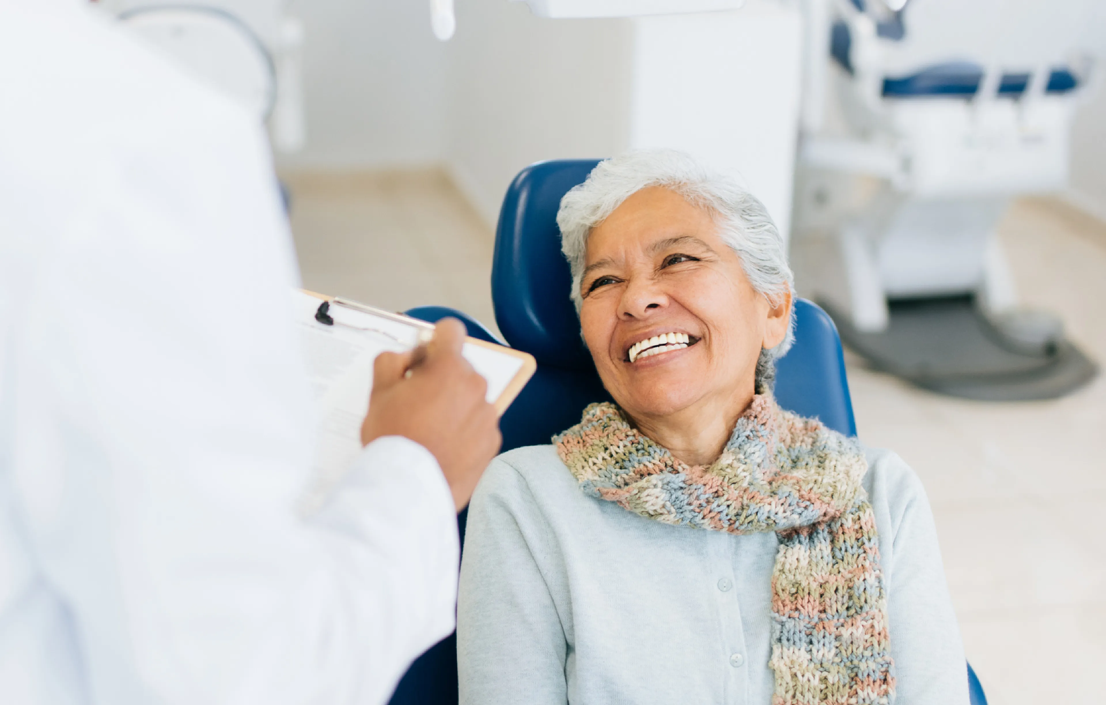An older woman wearing a scarf smiles while seated in a dental chair, speaking with a dentist holding a clipboard.